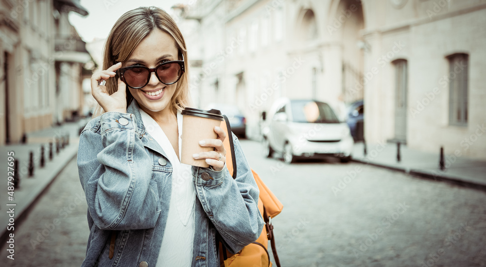 Fototapeta premium Nice beautiful millennial tourist with backpack in jeans jacket holds coffee cup and discover European city at holiday travel.