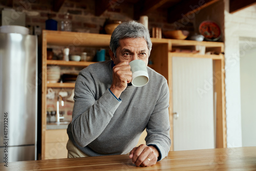 Portrait of retired biracial elderly male leaning on modern kitchen counter, sipping on his morning coffee.
