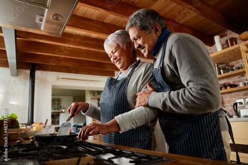 Elderly multi-ethnic couple smiling while cooking together in their modern kitchen. Happy and healthily retired