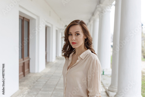pretty caucasian young woman in beige blouse, seductive red lipstick and wavy hair standing on veranda of cafe. white concrete pillars in background