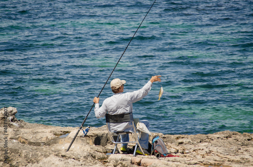 Up-Close of Successful Ocean Rock Fisherman Happily Reaching for a Fish He Caught While Angling Comfortably Sitting on a Chair Placed on a Rugged Rock Outcrop at the Edge of the Calm Blue Sea. Up-Close of Successful Ocean Rock Fisherman Happily Reaching for a Fish He Caught While Angling Comfortably Sitting on a Chair Placed on a Rugged Rock Outcrop at the Edge of the Calm Blue Sea.