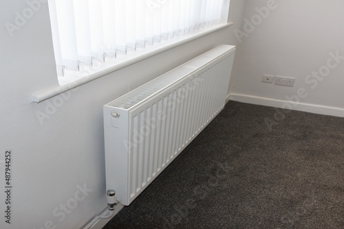 Corner of a room with a white radiator and white vertical blinds with a new carpet - house ready for rental or sale.
