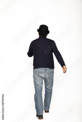 Man from behind walks. He is wearing a blue sweater, jeans and a bowler hat.