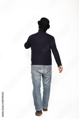 Man from behind walks. He is wearing a blue sweater, jeans and a bowler hat.