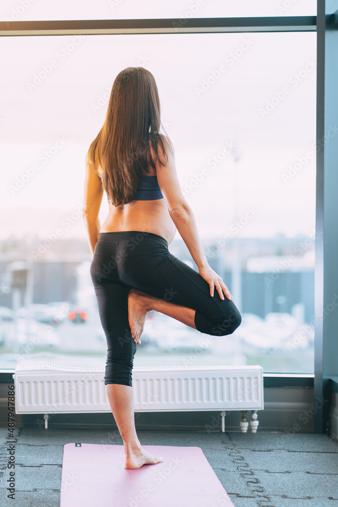 Vertical photo of slim athletic pregnant woman backside in uniform sportswear top, leggings. Doing yoga Tree Pose asana exercise, meditating in sport center near window, pink mat. Healthy sport