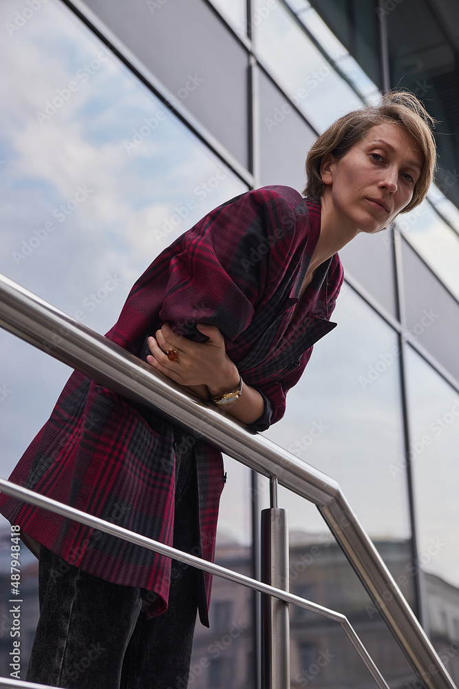 young woman with short hair in red checkered jacket outdoors in front of office building. portrait of caucasian female leaning on railings at street