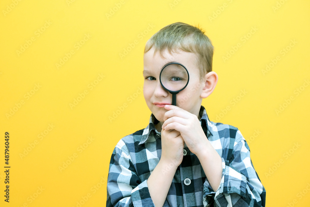 Positive curious schoolboy in casual clothes looks at the camera through a magnifying glass, on a yellow background. Positive curious schoolboy in casual clothes looks at the camera through a magnifying glass, on a yellow background.