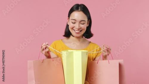 Charming young woman of Asian ethnicity 20s years old wear yellow t-shirt holding looking into package bags with purchases after shopping isolated on plain pastel light pink background studio portrait photo