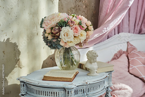 A bedside table in a room in the style of Provence or shabby chic. On the table there is a vase with flowers, a statuette and a book. Vintage interior.