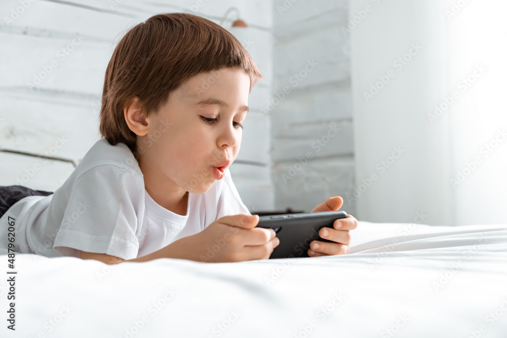 Little 5 year baby boy lays and plays with smartphone game. Wow emotions. Close-up child portrait. Holds in hands cell phone. Concept of online education. White room and clothes. Astonishment face Little 5 year baby boy lays and plays with smartphone game. Wow emotions. Close-up child portrait. Holds in hands cell phone. Concept of online education. White room and clothes. Astonishment face