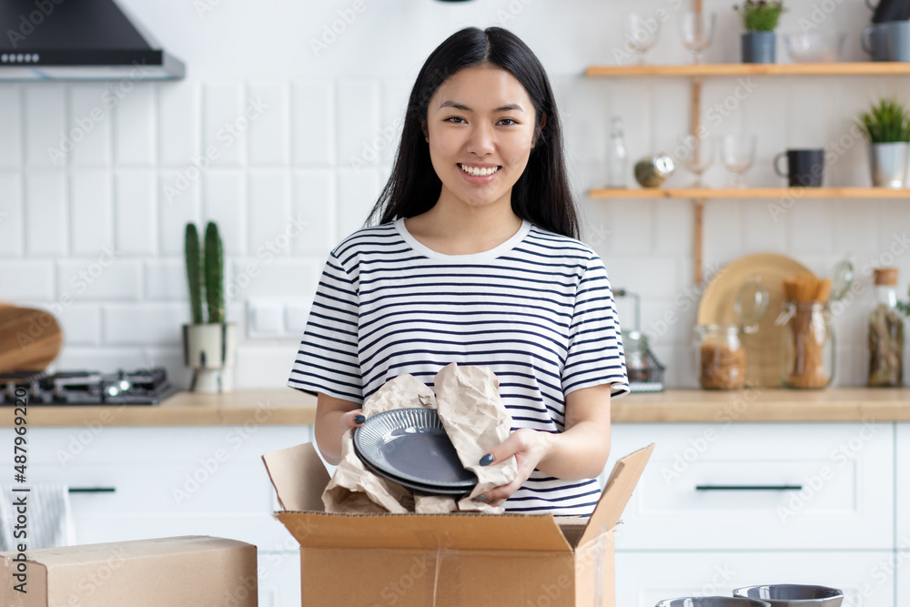 Young smiling Asian woman unpacking a box of purchased plates standing in the home kitchen looking at the camera and smiles friendly. Online shopping, delivery concept Young smiling Asian woman unpacking a box of purchased plates standing in the home kitchen looking at the camera and smiles friendly. Online shopping, delivery concept