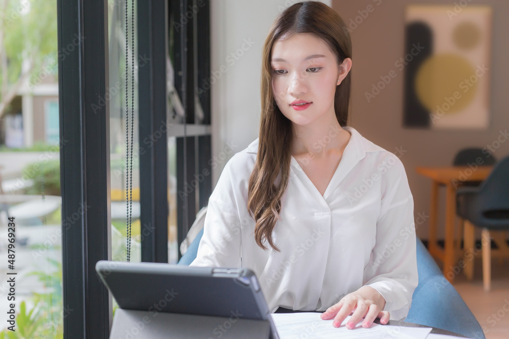 Asian confident woman wear a white shirt is using a tablet to work and there are many documents on the table. her face with smiling in a working room at home. Asian confident woman wear a white shirt is using a tablet to work and there are many documents on the table. her face with smiling in a working room at home.