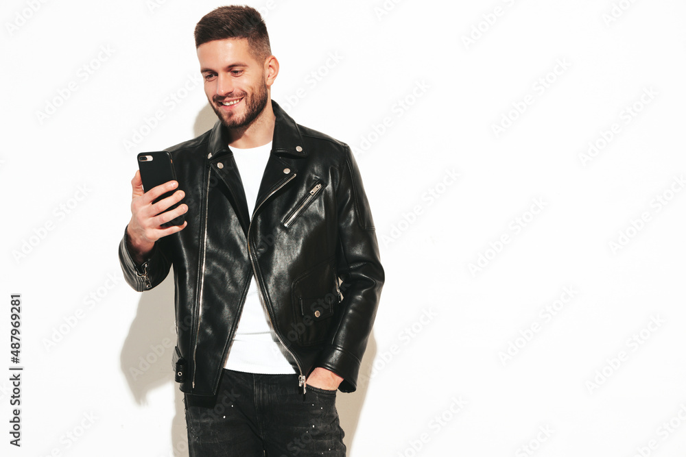 Portrait of handsome smiling model. Sexy stylish man dressed in biker leather jacket and black jeans. Fashion hipster male isolated on white in studio. Holding smartphone. Using apps Portrait of handsome smiling model. Sexy stylish man dressed in biker leather jacket and black jeans. Fashion hipster male isolated on white in studio. Holding smartphone. Using apps