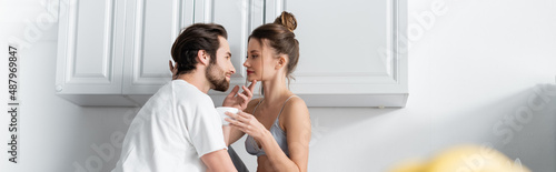bearded man seducing young woman in bra with cup in kitchen, banner.