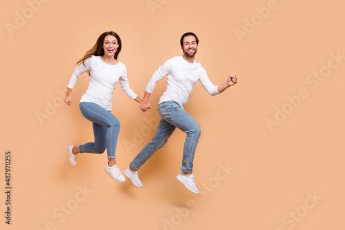Full length body size view of attractive cheerful partners jumping motion holding hands isolated over beige pastel color background