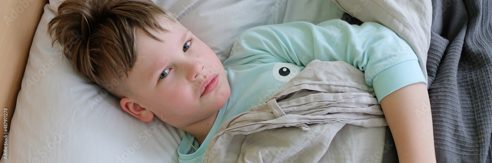 The child is lying with a fever in bed with a plush toy. A sad sick boy is lying in bed. Headache and high fever in case of flu or respiratory infection, virus. Epidemic of the disease. Banner for web The child is lying with a fever in bed with a plush toy. A sad sick boy is lying in bed. Headache and high fever in case of flu or respiratory infection, virus. Epidemic of the disease. Banner for web