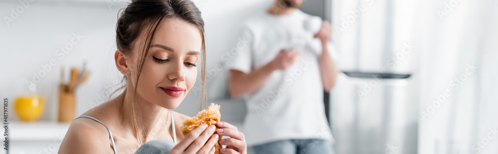 young woman holding croissant near blurred man on background, banner. young woman holding croissant near blurred man on background, banner.