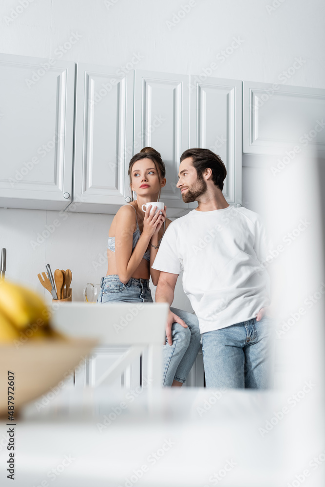 tattooed young woman in bra holding cup near bearded boyfriend in kitchen. tattooed young woman in bra holding cup near bearded boyfriend in kitchen.