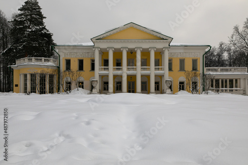 A vintage yellow manor house with white columns stands in a winter garden. It is surrounded by trees and a lot of snow. The drifts are deep and the trees are covered with snow.