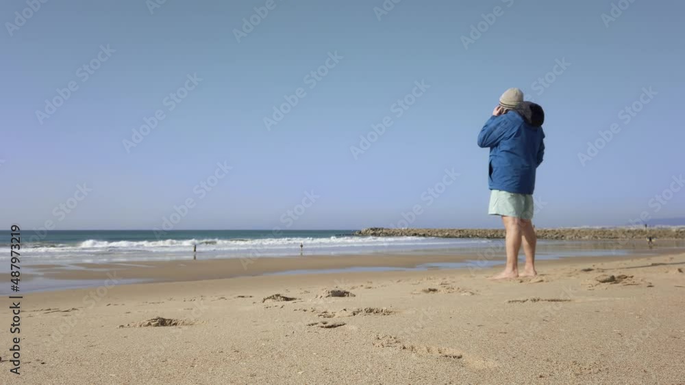 Older Male Taking Phone Call On Beach Wearing Blue Coat. Locked Off Older Male Taking Phone Call On Beach Wearing Blue Coat. Locked Off