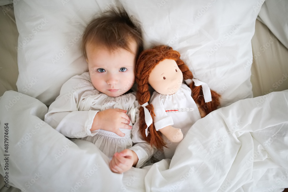 A cute Caucasian baby with a doll and with a thermometer, a sick child lies in bed with a toy, bed rest during illness. Comfort and play in bed A cute Caucasian baby with a doll and with a thermometer, a sick child lies in bed with a toy, bed rest during illness. Comfort and play in bed