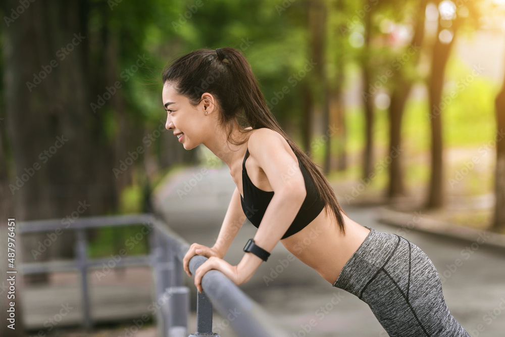 Naklejka premium Asian fitness girl doing push-ups for chest and arms with handrail outdoors
