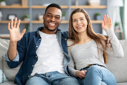 Portrait of happy multicultural couple sitting on couch, waving hands at camera