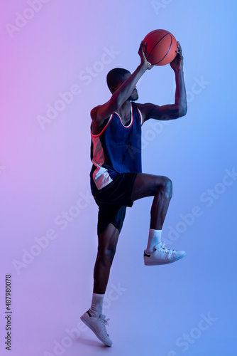 Full length portrait of professional black basketball player jumping and doing scoring shot in neon light © Prostock-studio