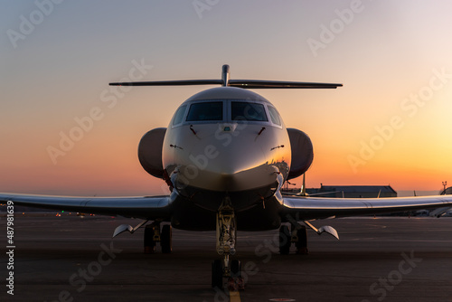 Scenic front view modern luxury expensive private jet plane parked airport taxiway hangar warm colorful dramatic evening warm sunset sun light sky background. Executive aicraft vip travel concept