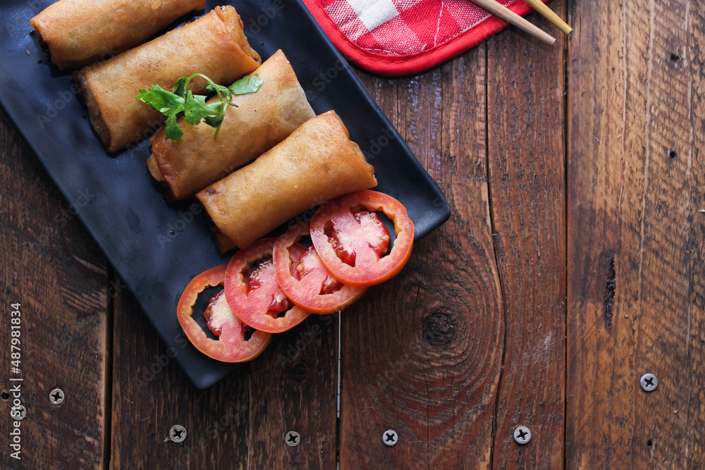 Fried spring rolls served in a black square plate with sliced tomatoes on a brown wooden table.