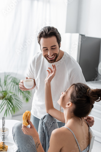 young woman holding fresh croissant and reaching happy boyfriend with cup.