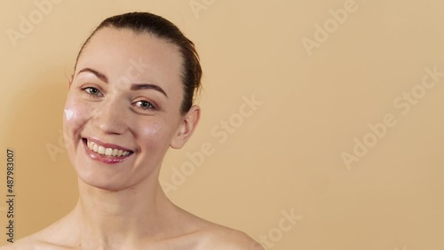 Attractive caucasian girl applying a cosmetic cream on her face on her cheek isolated on a beige studio background. Skin care concept.