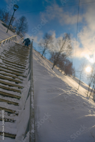 Street photo with a steep staircase and a man climbing it. A sunny winter day. Blinding winter sun, snow-covered hill and steep stairs up. Original angle