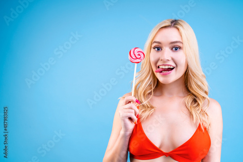 Closeup portrait of young blond woman in red swimsuit on blue background. Happy and joyful girl is holding bright colorful lollipop sweets candies in hands. Vacation on sea beach concept. Summer mood