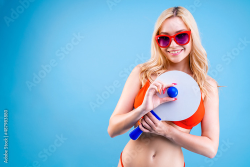 Young sunny beautiful woman in red swimsuit and glasses ready to play beach games. Blond smiling girl is holding matkot racket and ball against blue background. Summer mood. Sea vacation concept. photo