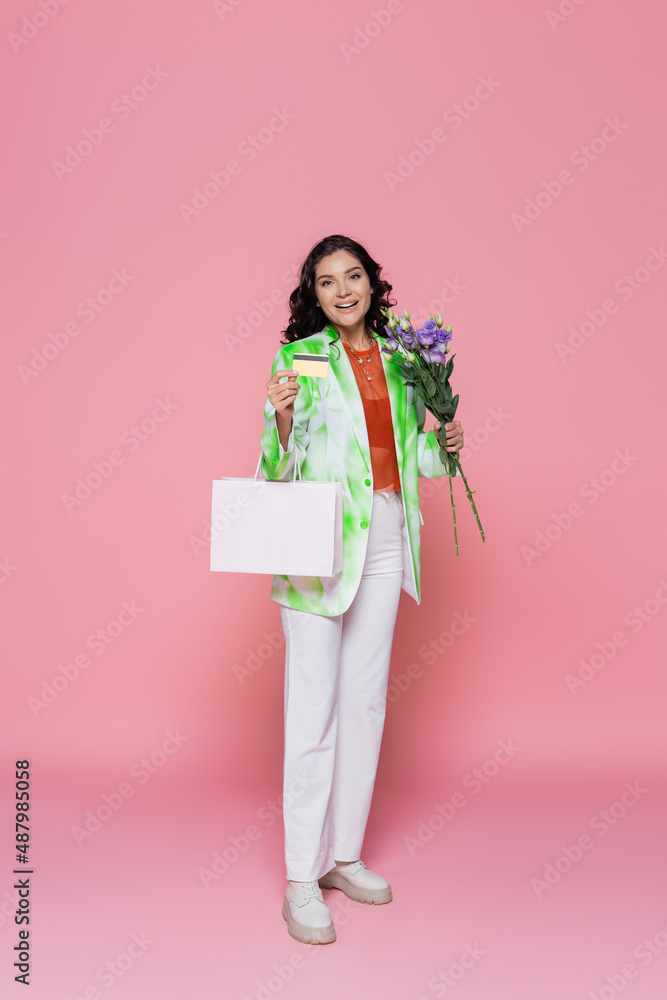 full length of smiling young woman in tie dye blazer holding credit card, shopping bag and flowers on pink. full length of smiling young woman in tie dye blazer holding credit card, shopping bag and flowers on pink.