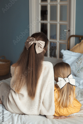 Portrait from behind of loving beautiful young mother and little daughter with long gorgeous hair with a trendy hairpin bow at home on bedroom sitting in cozy bed. Happy mothers day