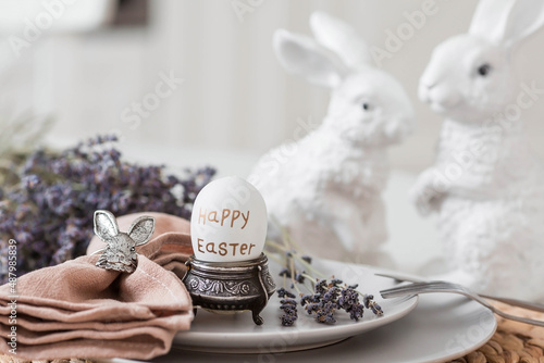 Table setting. A plate of Easter eggs, rabbits, lavender and a napkin with appliances on a white table. There is a Scandinavian-style kitchen in the background. The concept of a bright Easter holiday.