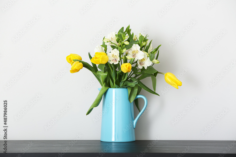 Jug with bouquet of flowers on table near light wall