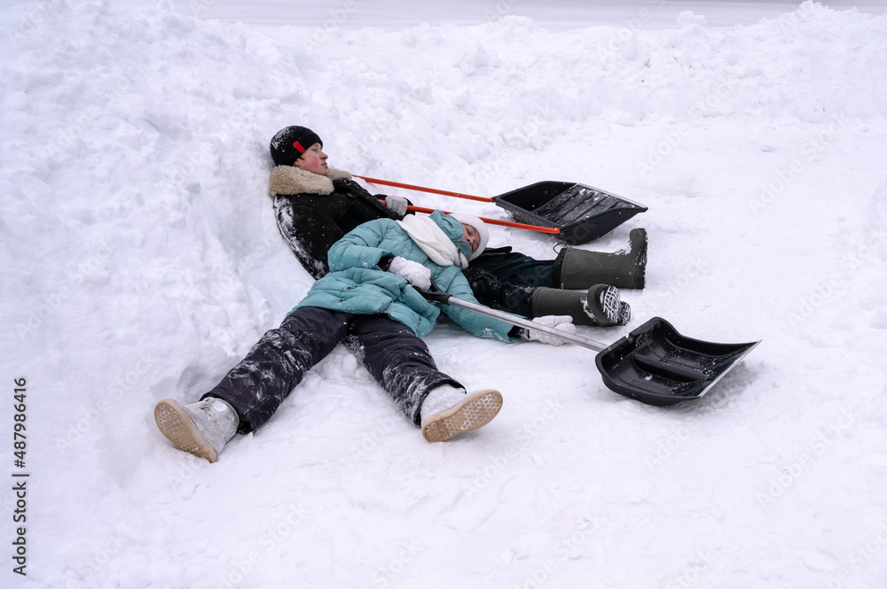 girl and guy in winter clothes are tired and lie resting in  snow in winter. There are big shovels next to them. Rest after snow removal