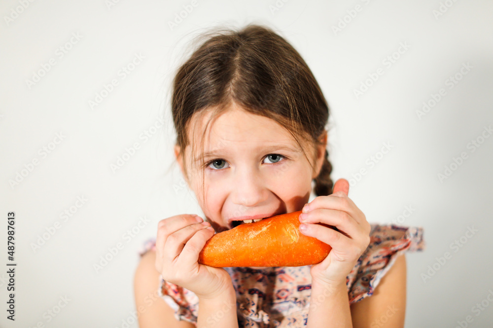 Funny caucasian girl child with carrots on a light background, vegetables and vitamins for children. Strong kid nibbles on a carrot Funny caucasian girl child with carrots on a light background, vegetables and vitamins for children. Strong kid nibbles on a carrot