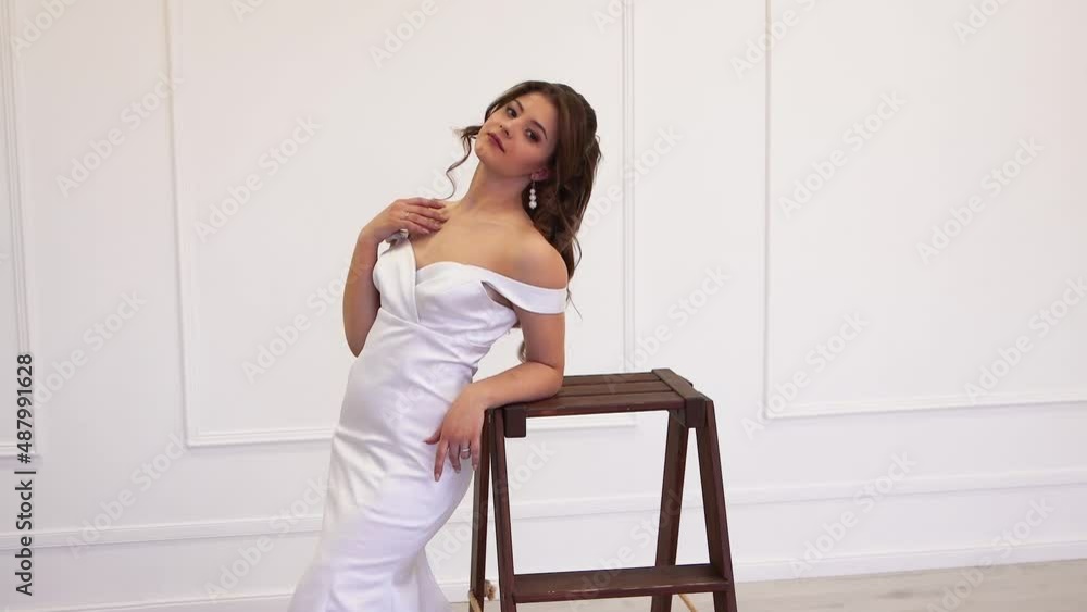 Beautiful bride-model in a white dress poses for a photographer. Gorgeous modern girl on a light isolated background in the studio. Warm soft light, copy space. UHD 4K. Beautiful bride-model in a white dress poses for a photographer. Gorgeous modern girl on a light isolated background in the studio. Warm soft light, copy space. UHD 4K.