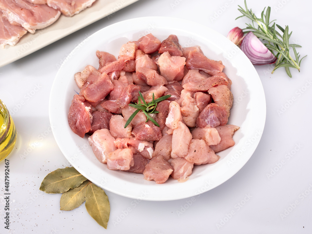 Diced meat on a white plate. Composition with raw poultry, pork meat in cubes and seasonings, on white background.