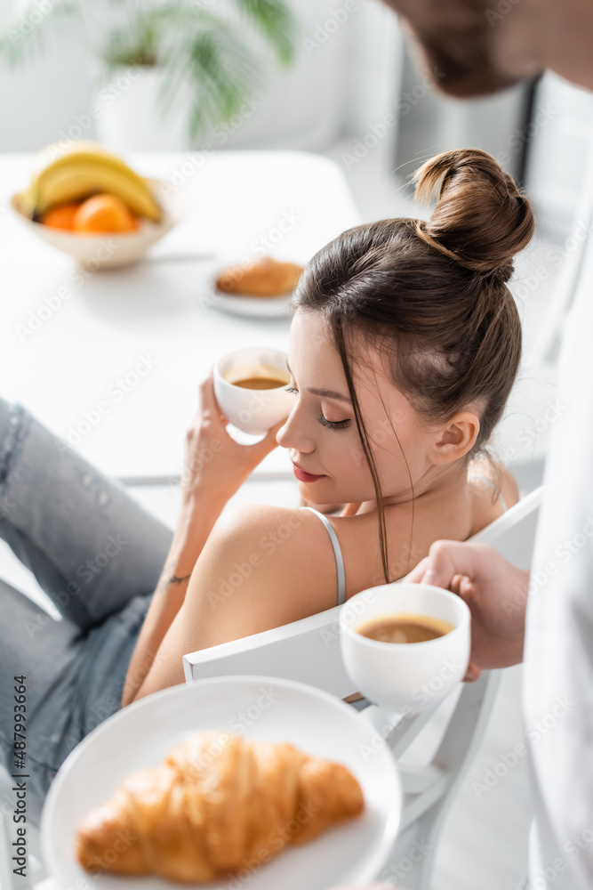 tattooed woman holding cup near blurred man with croissant on plate.