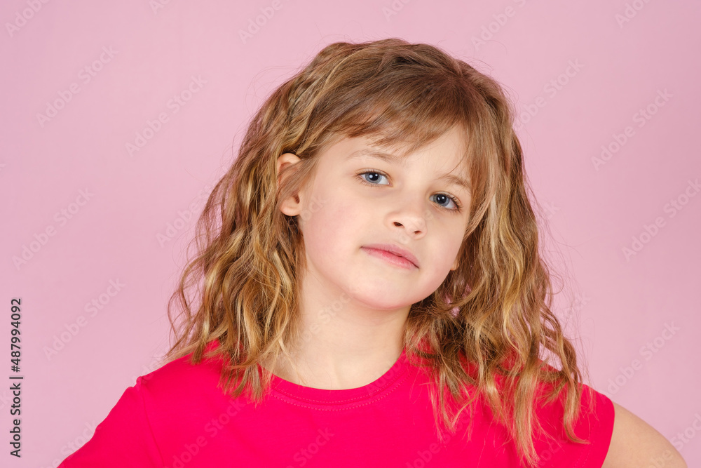 Close-up adorable little girl with long curly hair in red colored t-shirt looking at camera, against pink background in studio Close-up adorable little girl with long curly hair in red colored t-shirt looking at camera, against pink background in studio