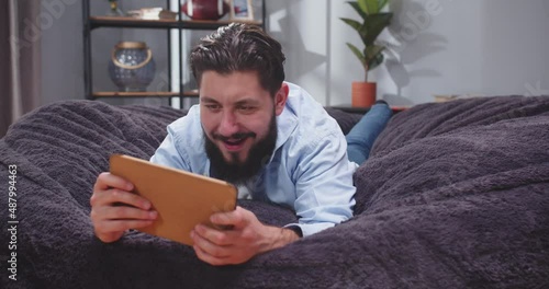 Close up portrait of adult man lying on his stomach on sofa and playning game on tablet. Male scrolling social media on gadget. Man after work resting on sofa with tablet. Slow motion video. photo