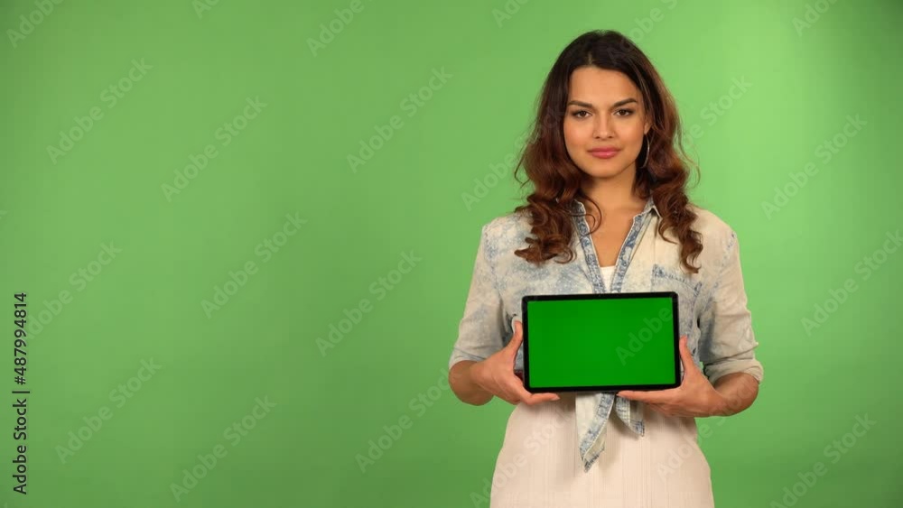 A young beautiful Caucasian woman shows a tablet with green screen to the camera with a smile - green screen background A young beautiful Caucasian woman shows a tablet with green screen to the camera with a smile - green screen background