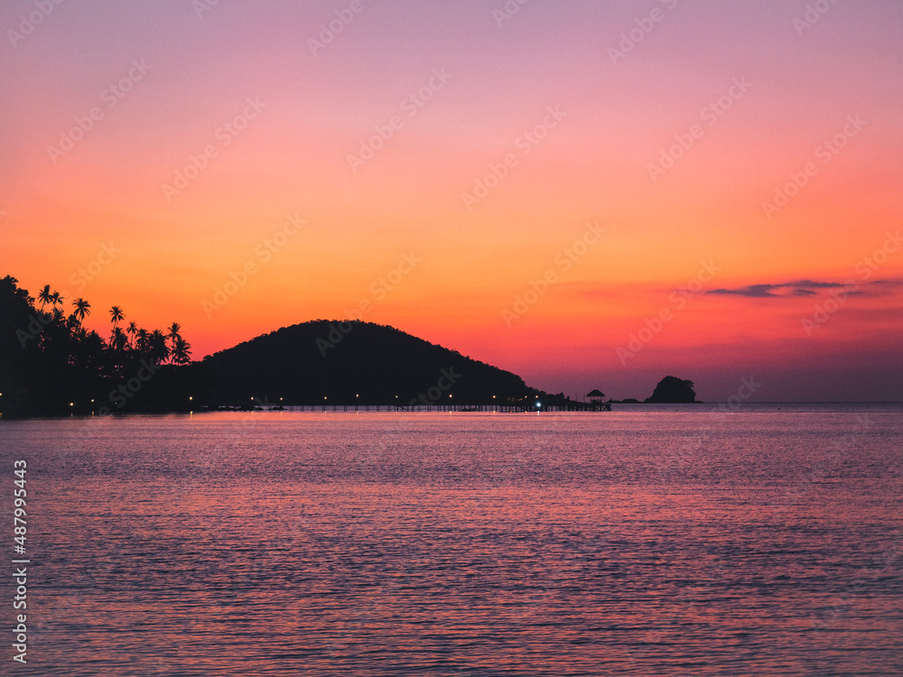 Scenic view of colorful sunset sky at peaceful sea bay with long wooden pier landmark and island silhouette. Koh Mak Island, Trat Province, Thailand.