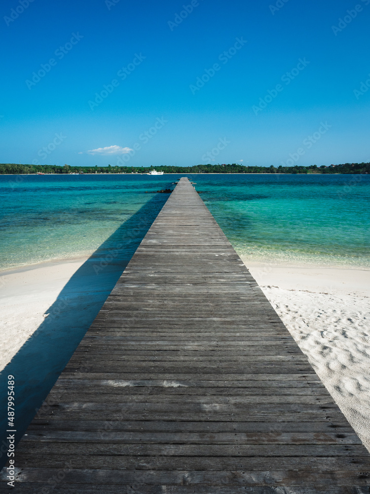 Long straight wooden pier over crystal clear turquoise water and white sand beach of Koh Kham Island. Look to Koh Mak Island, Trat Province, Thailand.