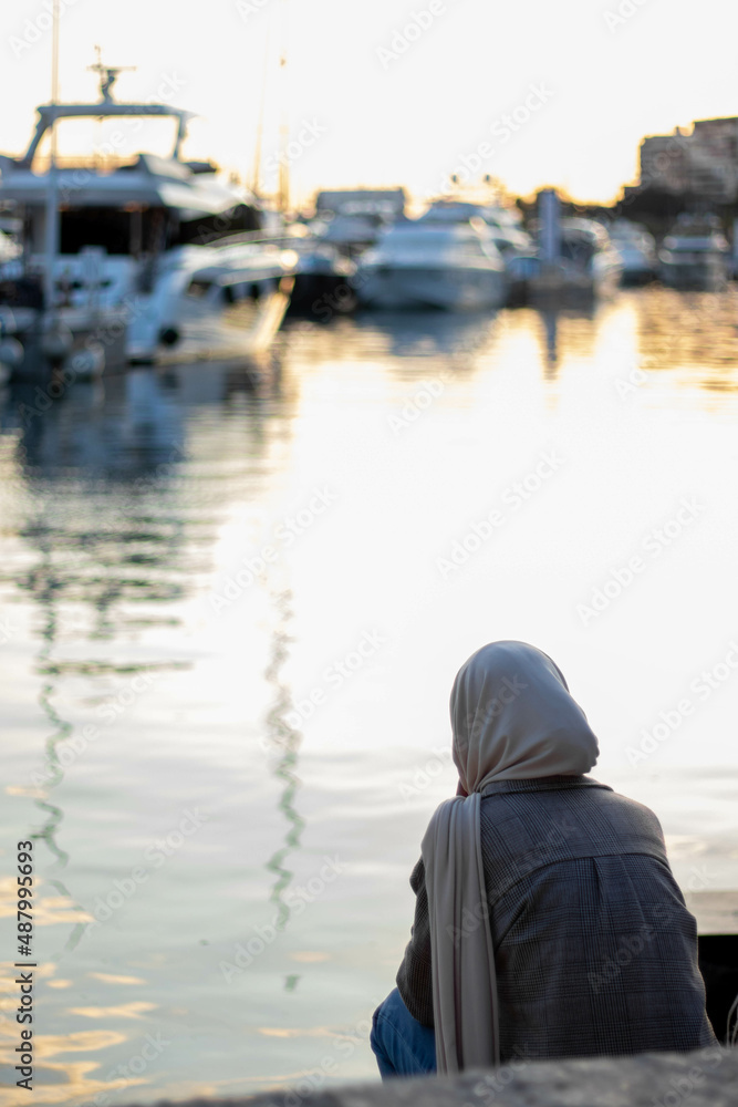 Fototapeta premium Muslim woman with hijab on her back facing the sea in the harbor